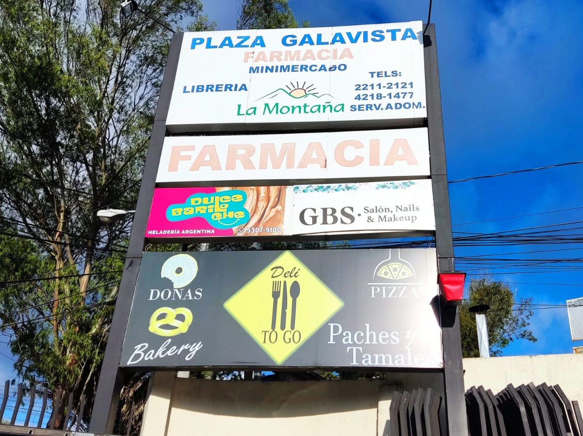 Tótem exterior iluminado con tiendas como una farmacia, un salón de belleza y una pizzería.